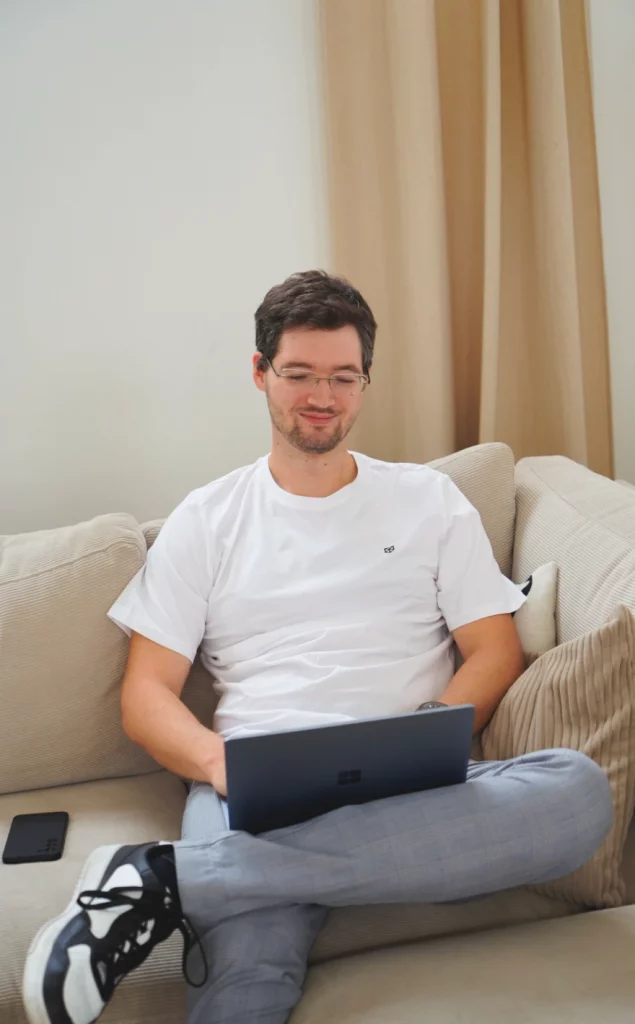 Junger Mann in heller Kleidung auf einer cremefarbenen Couch arbeitet mit seinem Laptop.